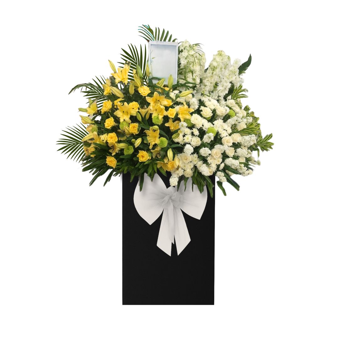 Floral condolences arrangement with yellow and white flowers on a black stand with a white bow, against a white background.