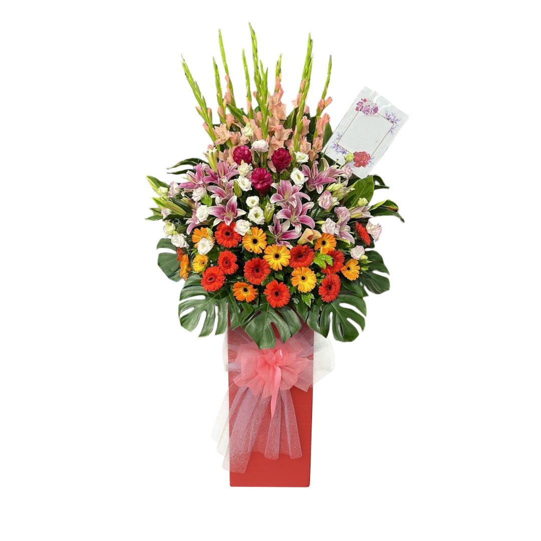 Vibrant grand opening flower stand with orange, red, and yellow gerberas, pink lilies, and tall snapdragons in a red box base, commonly used to celebrate new business openings or achievements.