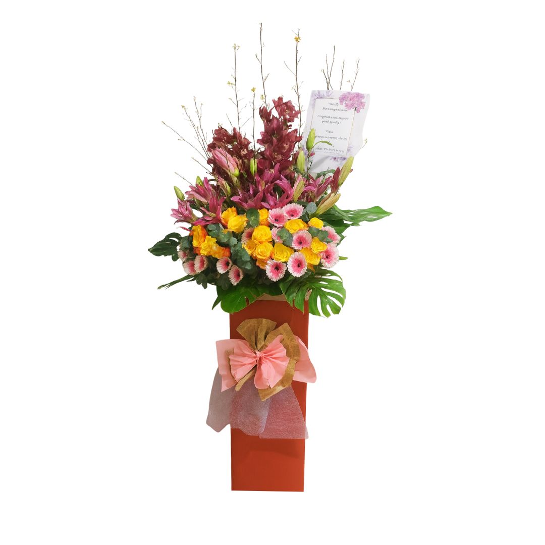 Tall grand opening flower stand with burgundy and pink lilies, yellow and pink gerberas, and large tropical leaves in a tall, red base with a large pink bow, commonly used to celebrate new business openings or achievements.
