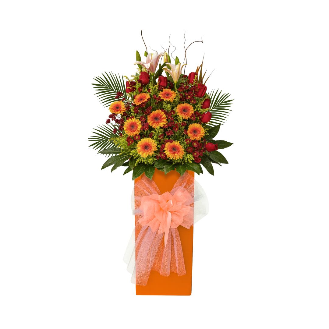 Vibrant grand opening flower stand with orange gerbera daisies, red roses, and lilies in a tall orange base, ideal for business celebrations.