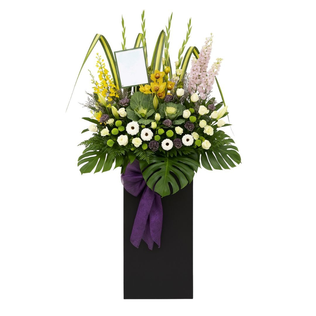Floral condolences arrangement in a black container with a purple ribbon on a white background