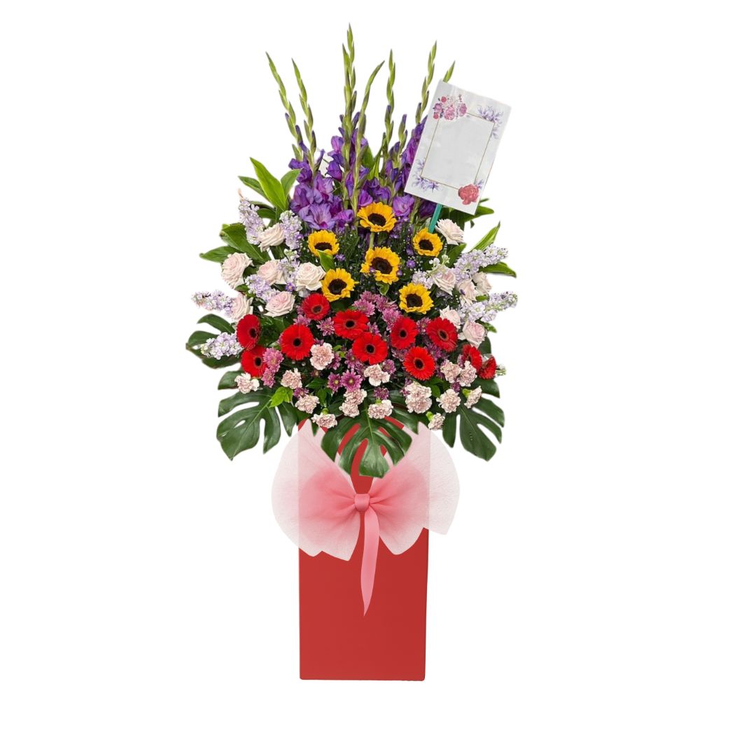 Vibrant grand opening flower stand with sunflowers, red gerberas, and mixed white and purple flowers in a tall red box with a pink bow, designed for a business launch or celebration.
