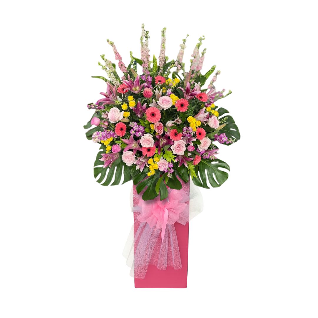Tall grand opening flower stand with pink lilies, pink and yellow gerberas, light pink roses, purple snapdragons, and monstera leaves in a pink stand with a tulle bow, commonly sent to celebrate new business openings.