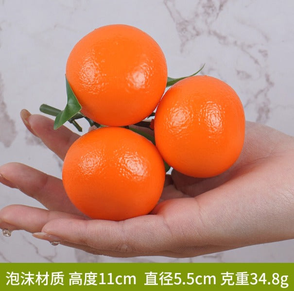 Three artificial oranges clustered on a green stem held in a person's hand. These are lifelike foam or plastic decorative fruit models often used for home decor, displays, or photography props.
