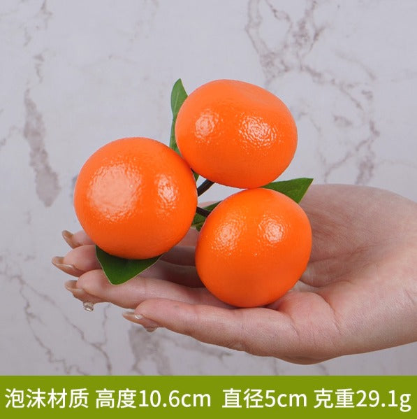 Three artificial oranges clustered on a green stem held in a person's hand. These are lifelike foam or plastic decorative fruit models often used for home decor, displays, or photography props.